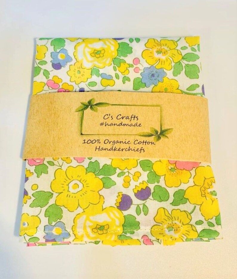 May include: A white cotton handkerchief with a yellow, pink, and blue floral pattern. The handkerchief is folded and has a brown paper label that reads "C's Crafts #handmade 100% Organic Cotton Handkerchiefs".