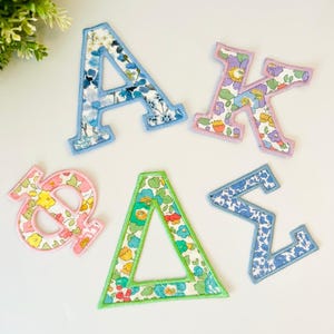 May include: Five fabric patches shaped as Greek letters. The letter A has a blue floral pattern with light blue stitching. The letter K features a purple, yellow, and blue floral print with pink stitching. The Phi has pink stitching and a yellow floral pattern. The Delta has green stitching and a teal floral print. The Sigma has blue stitching and a blue floral pattern.