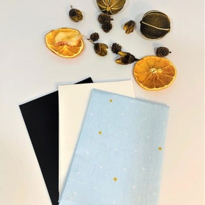 May include: Three fabric squares, one black, one white, and one light blue with white dots and gold stars. The fabric squares are arranged on a white surface with dried orange slices and other natural elements.