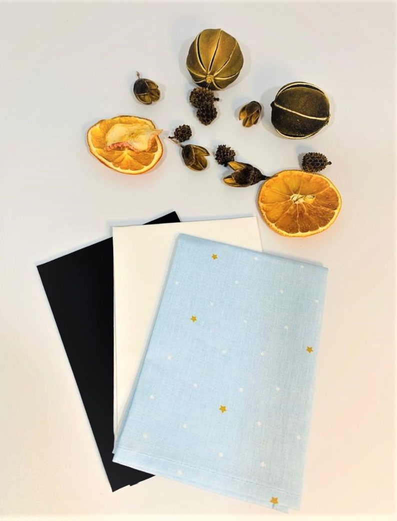 May include: Three fabric squares, one black, one white, and one light blue with white dots and gold stars. The fabric squares are arranged on a white surface with dried orange slices and other natural elements.