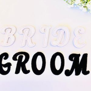 Könnte beinhalten: Weiße Filzbuchstaben, die "BRIDE" buchstabieren, und schwarze Filzbuchstaben, die "GROOM" buchstabieren, auf weißem Hintergrund.