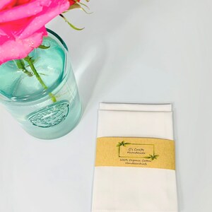 May include: Two white cotton handkerchiefs folded and stacked on a white surface. A brown label with the text "C's Crafts #handmade 100% Organic Cotton Handkerchiefs" is attached to the handkerchiefs.