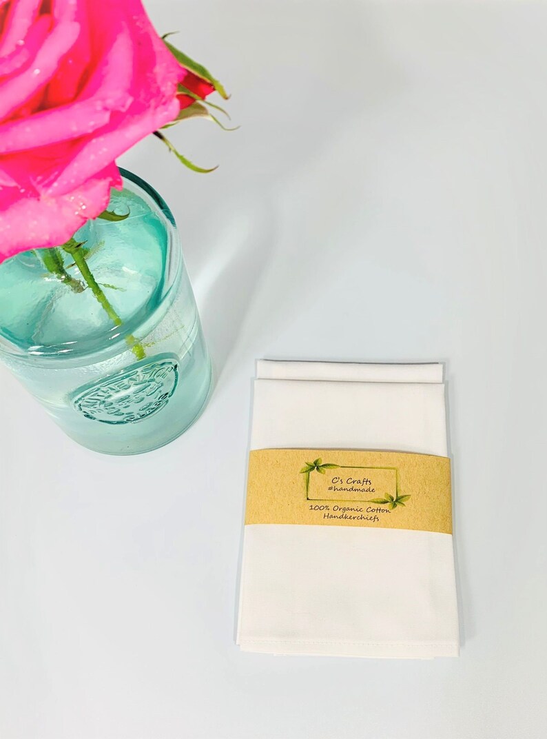 May include: Two white cotton handkerchiefs folded and stacked on a white surface. A brown label with the text "C's Crafts #handmade 100% Organic Cotton Handkerchiefs" is attached to the handkerchiefs.