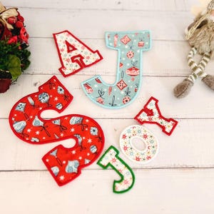 May include: Fabric letters spelling out AJSOY with various holiday patterns including red, light blue, and white colours. The letters feature festive motifs like ornaments and stars, with stitched edges, arranged on a white wooden surface.