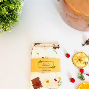 May include: Two white cotton handkerchiefs with a brown book and floral print. The handkerchiefs are folded and have a yellow label that reads "C's Crafts #handmade 100% Cotton Handkerchiefs".