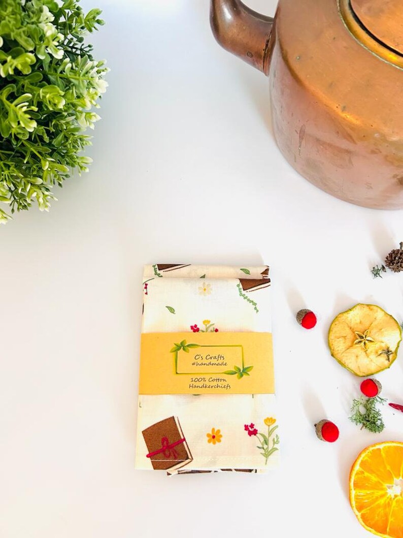 May include: Two white cotton handkerchiefs with a brown book and floral print. The handkerchiefs are folded and have a yellow label that reads "C's Crafts #handmade 100% Cotton Handkerchiefs".