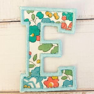 May include: A fabric letter E patch featuring a floral pattern with teal, green, yellow, and coral flowers on a white background, finished with a light blue embroidered satin stitch border around the entire edge of the letter shape.