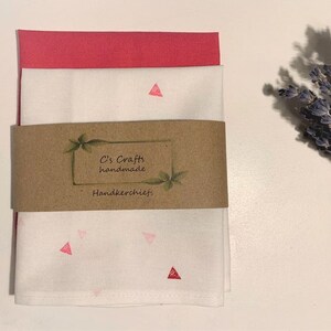 May include: Two white handkerchiefs with pink triangle patterns. The top handkerchief is folded and the bottom handkerchief is unfolded. A brown paper tag with the text "C's Crafts handmade Handkerchiefs" is attached to the handkerchiefs.