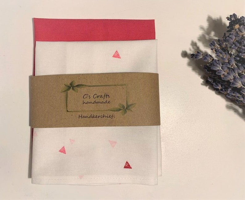 May include: Two white handkerchiefs with pink triangle patterns. The top handkerchief is folded and the bottom handkerchief is unfolded. A brown paper tag with the text "C's Crafts handmade Handkerchiefs" is attached to the handkerchiefs.