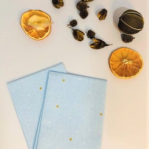 May include: Two light blue tea towels with a white polka dot pattern and gold stars. The towels are folded and stacked on a white surface.