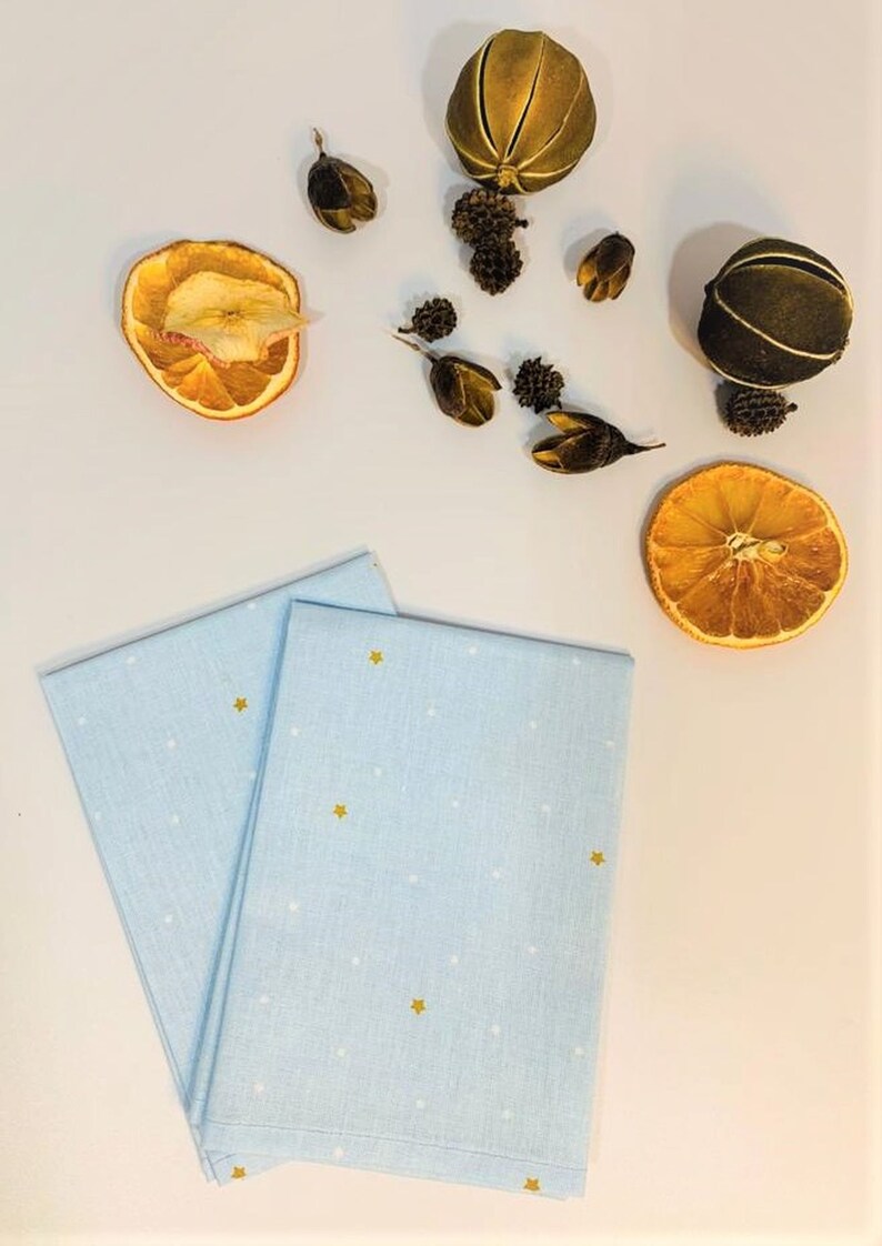May include: Two light blue tea towels with a white polka dot pattern and gold stars. The towels are folded and stacked on a white surface.