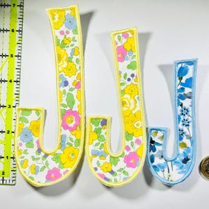 May include: Three fabric letter J patches with floral patterns. Two patches feature yellow, pink, and blue flowers with yellow borders, while the third has blue flowers with a light blue border. A yellow ruler shows the height is 15 cm.