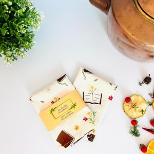 May include: Two white cotton handkerchiefs with a floral and book design. The handkerchiefs are folded and stacked on top of each other. A small yellow tag with the text "C's Crafts #handmade 100% Cotton Handkerchiefs" is attached to the handkerchiefs.