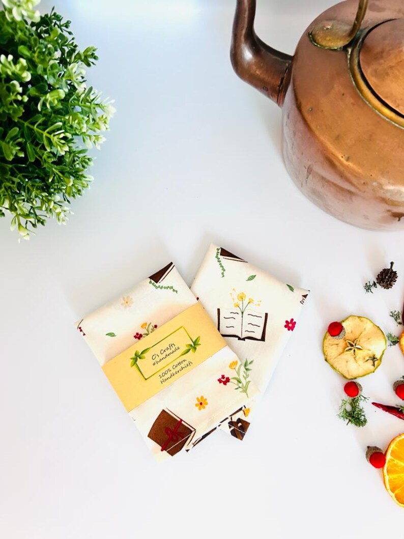 May include: Two white cotton handkerchiefs with a floral and book design. The handkerchiefs are folded and stacked on top of each other. A small yellow tag with the text "C's Crafts #handmade 100% Cotton Handkerchiefs" is attached to the handkerchiefs.