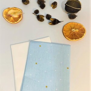 May include: Two fabric napkins, one white and one light blue with white dots and gold stars. The napkins are folded and arranged on a white surface. Dried orange slices and other natural elements are scattered around the napkins.