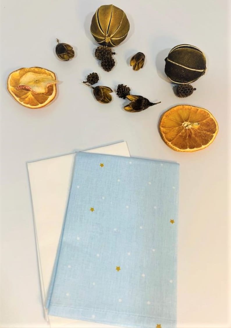 May include: Two fabric napkins, one white and one light blue with white dots and gold stars. The napkins are folded and arranged on a white surface. Dried orange slices and other natural elements are scattered around the napkins.