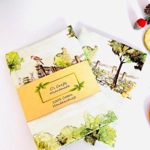 May include: Two white cotton handkerchiefs with a watercolor print of a countryside scene. The handkerchiefs are folded and stacked on top of each other. A yellow tag with the text "C's Crafts #handmade 100% Cotton Handkerchiefs" is attached to the top handkerchief.