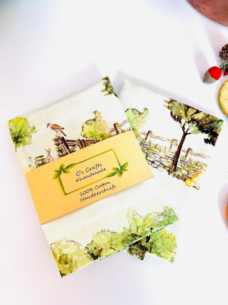 May include: Two white cotton handkerchiefs with a watercolor print of a countryside scene. The handkerchiefs are folded and stacked on top of each other. A yellow tag with the text "C's Crafts #handmade 100% Cotton Handkerchiefs" is attached to the top handkerchief.