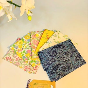 Op de afbeelding: Een set van zeven stoffen vierkanten, waaronder een blauwe paisley print, een gele bloemenprint en vijf bloemenprints in tinten roze, geel en blauw. De stoffen vierkanten zijn in een waaier vorm op een wit oppervlak gerangschikt. Een klein bruin label met de tekst "C's Crafts #handmade" bevindt zich in de rechter benedenhoek van de afbeelding.