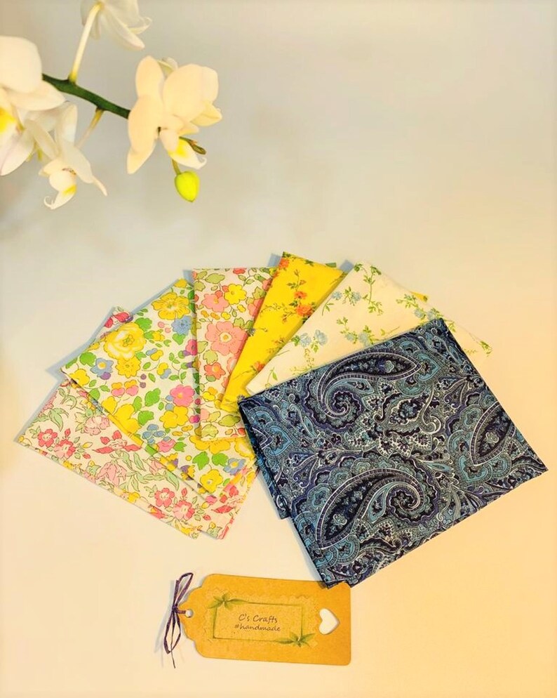 Op de afbeelding: Een set van zeven stoffen vierkanten, waaronder een blauwe paisley print, een gele bloemenprint en vijf bloemenprints in tinten roze, geel en blauw. De stoffen vierkanten zijn in een waaier vorm op een wit oppervlak gerangschikt. Een klein bruin label met de tekst "C's Crafts #handmade" bevindt zich in de rechter benedenhoek van de afbeelding.