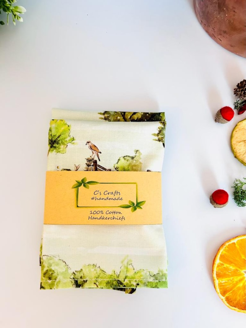 May include: A folded white cotton handkerchief with a green and brown watercolor design of a bird perched on a branch. The handkerchief has a label that reads "C's Crafts #handmade 100% Cotton Handkerchiefs".