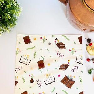 May include: A white tea towel with a brown and red book pattern. The books are surrounded by small flowers and leaves. The tea towel is on a white surface.