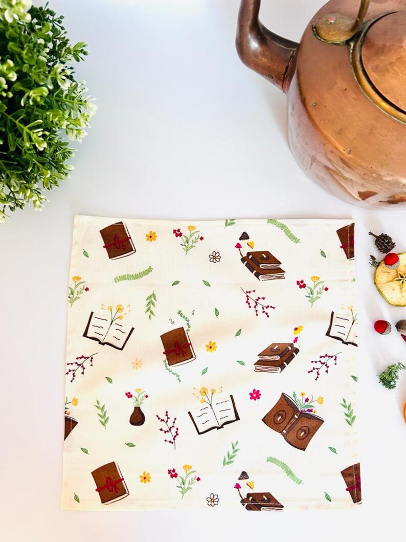 May include: A white tea towel with a brown and red book pattern. The books are surrounded by small flowers and leaves. The tea towel is on a white surface.