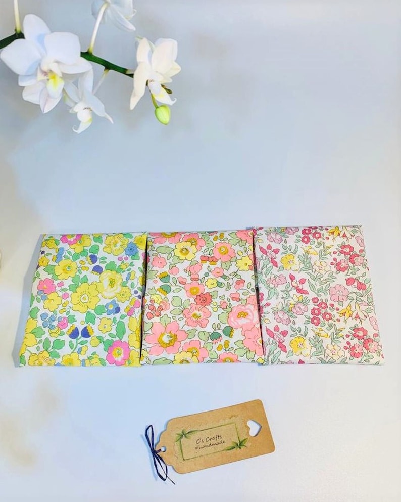 May include: Three small fabric pouches with floral patterns. The pouches are lined up in a row on a white surface. The pouch on the left has a yellow floral pattern, the middle pouch has a pink floral pattern, and the pouch on the right has a pink and white floral pattern. A small brown tag with the text "C's Crafts #handmade" is in the lower right corner of the image.