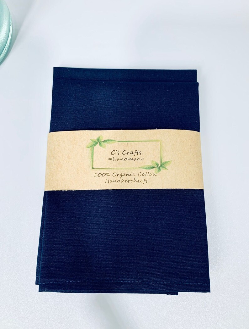 May include: A stack of three dark blue 100% organic cotton handkerchiefs with a brown paper label that reads "C's Crafts #handmade 100% Organic Cotton Handkerchiefs".
