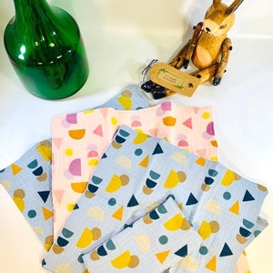 May include: A stack of four blue and pink fabric squares with geometric patterns. The squares are made of a soft, textured material and feature a variety of shapes, including circles, triangles, and semi-circles. The squares are arranged in a staggered pattern, with the largest square at the bottom and the smallest square at the top.