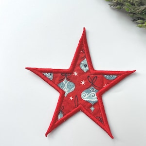 May include: A star-shaped fabric appliqué with a red background featuring a repeating pattern of light blue and white Christmas ornaments and small white stars. The edges of the star are finished with a thick, red embroidered satin stitch border.