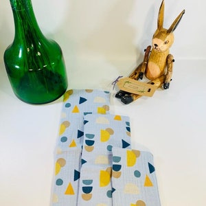 May include: A set of four blue fabric cloths with a geometric pattern of yellow, brown, and blue circles, triangles, and semi-circles. The cloths are folded and stacked on a white surface.