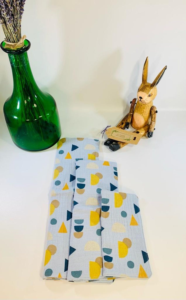 May include: A set of four blue fabric cloths with a geometric pattern of yellow, brown, and blue circles, triangles, and semi-circles. The cloths are folded and stacked on a white surface.