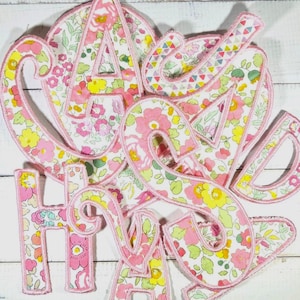 May include: Fabric letters spelling "HAVE A DREAM" in a floral pattern with pink and yellow flowers on a white background. Each letter is outlined in pink and has a stitched edge. The letters are arranged in a decorative manner.