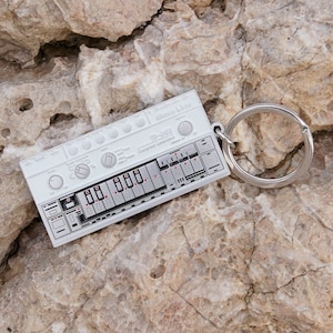 Peut inclure: Un porte-clés blanc avec un design de synthétiseur miniature. Le porte-clés est orné d'un anneau argenté et porte l'inscription "TB-303" et "Bass Line". Le synthétiseur présente des détails noirs et rouges.