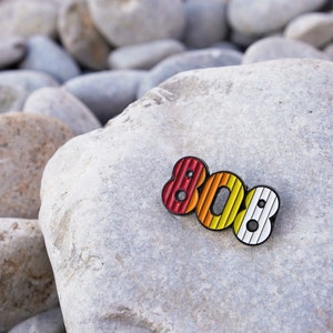 Puede incluir: Un pin de esmalte colorido con el número 808 en un diseño a rayas. El pin es rojo, naranja y blanco. El pin está sobre un fondo de roca gris.