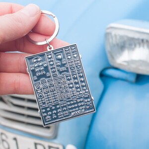 Puede incluir: Un llavero de metal plateado con un diseño detallado de un panel de control de sintetizador. El llavero está unido a un coche azul.