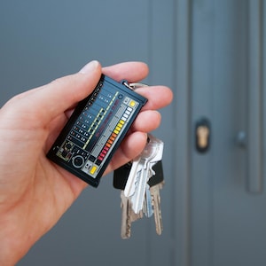 Puede incluir: Un llavero negro y rojo con forma de sintetizador vintage con el texto "Roland Compact TR-606" en la parte delantera. El llavero está unido a un juego de llaves.