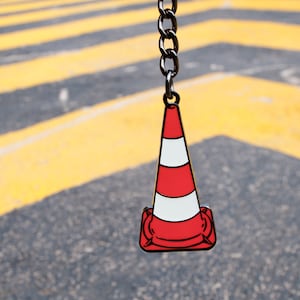 Puede incluir: Un llavero de cono de tráfico rojo y blanco con una cadena plateada. El cono es de metal y tiene un diseño detallado.