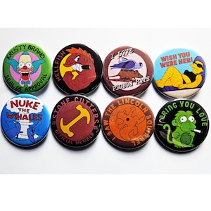 May include: A collection of eight round pinback buttons with various cartoon character designs and text. The buttons feature characters and phrases like "Krusty Brand Seal of Approval," "Nuke the Whales," and "I Love Pigeon Rats."