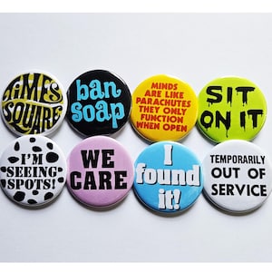 Op de afbeelding: Een verzameling van acht ronde buttons met verschillende tekstontwerpen. De buttons tonen zinnen als "Times Square", "ban soap" en "I'm seeing spots!" in vetgedrukte, kleurrijke lettertypen. De buttons staan op een witte achtergrond.