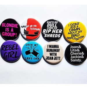 Könnte beinhalten: Acht runde Anstecknadeln mit verschiedenen Slogans und Bandnamen. Die Nadeln sind schwarz, weiß, gelb, rot, blau und lila. Die Slogans lauten: "Blondie is a group!" "Rebel Girl" "She's so dull come on rip her to shreds" "I don't give a damn about my bad reputation" "Hanging on the telephone" "Suck my left one" "I wanna runaway with Joan Jett" "Joan & Lita & Cherie & Jackie & Sandy."