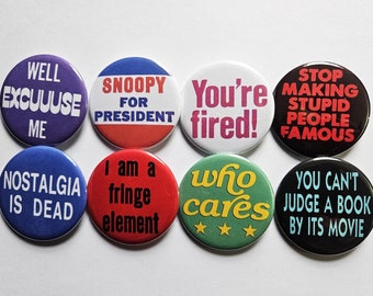 Puede incluir: Ocho pines redondos con diferentes lemas humorísticos. Los pines son negros, rojos, azules, verdes y blancos con texto negro. Los lemas incluyen "Well Excuse Me", "Snoopy for President", "You're Fired!", "Stop Making Stupid People Famous", "Nostalgia is Dead", "I am a Fringe Element", "Who Cares", y "You Can't Judge a Book by Its Movie".