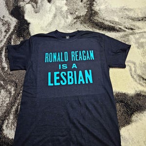 May include: A black short-sleeved t-shirt featuring the text "RONALD REAGAN IS A LESBIAN" printed in bright cyan blue lettering across the chest. The shirt has a crew neckline and a standard fit, with the graphic centered on the front panel.