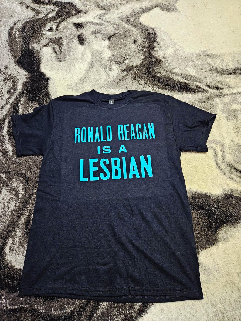 May include: A black short-sleeved t-shirt featuring the text "RONALD REAGAN IS A LESBIAN" printed in bright cyan blue lettering across the chest. The shirt has a crew neckline and a standard fit, with the graphic centered on the front panel.