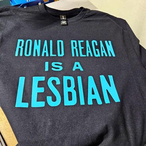 May include: A black short-sleeved t-shirt featuring bright cyan text printed on the front. The text reads RONALD REAGAN IS A LESBIAN in a bold, sans-serif font. The garment has a crew neckline and a black fabric label at the inner collar.