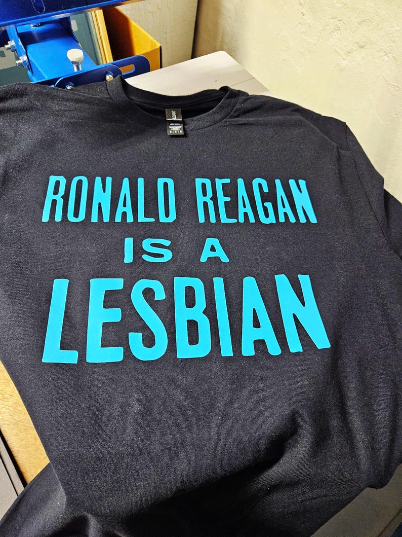 May include: A black short-sleeved t-shirt featuring bright cyan text printed on the front. The text reads RONALD REAGAN IS A LESBIAN in a bold, sans-serif font. The garment has a crew neckline and a black fabric label at the inner collar.