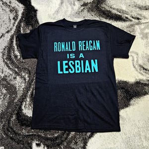 May include: A black short-sleeved t-shirt featuring the text RONALD REAGAN IS A LESBIAN printed in a bright cyan blue font across the chest. The garment has a crew neckline and a standard fit, with the bold lettering centered on the front panel.