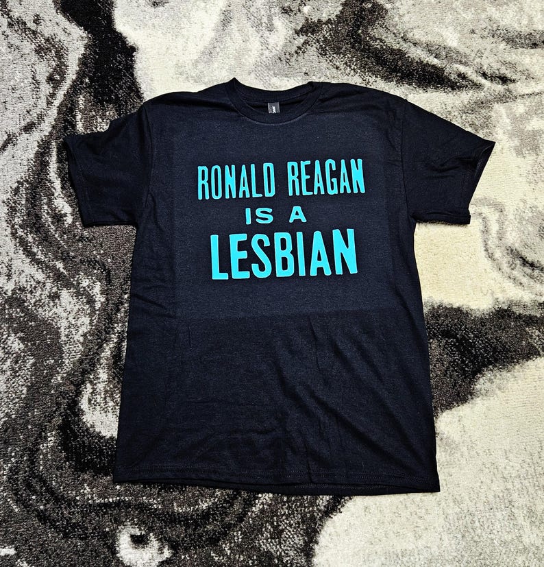 May include: A black short-sleeved t-shirt featuring the text RONALD REAGAN IS A LESBIAN printed in a bright cyan blue font across the chest. The garment has a crew neckline and a standard fit, with the bold lettering centered on the front panel.