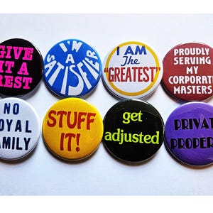 Puede incluir: Una colección de ocho botones redondos con varias frases en texto en negrita. Los botones son negros, azules, blancos, amarillos, rojos y morados. Las frases incluyen "GIVE IT A REST" y "I AM The GREATEST".